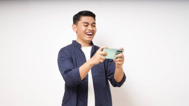 Heyecanlı Asyalı genç adam beyaz arka planda akıllı telefondan oyun oynuyor.