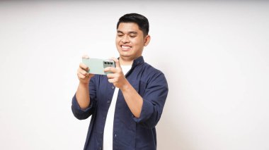 Heyecanlı Asyalı genç adam beyaz arka planda akıllı telefondan oyun oynuyor.