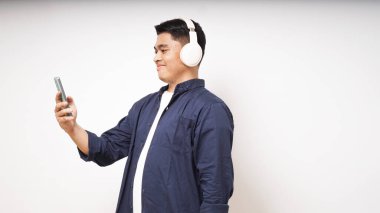 Beyaz arka planda izole edilmiş akıllı telefon stüdyosundan müzik dinleyen, kablosuz kulaklık takan mutlu Asyalı genç adam.