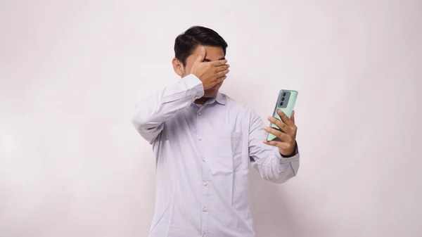 Asian man covering his eyes while looking to his mobile phone with scared expression against white background