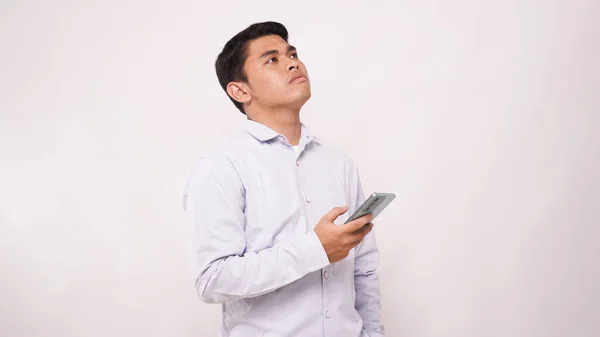 Adult Asian man thinking about something while holding mobile phone against white background