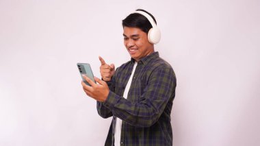 Akıllı telefondan müzik dinleyen Asyalı genç adam beyaz arka planda kablosuz kulaklık kullanıyor.