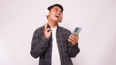Akıllı telefondan müzik dinleyen Asyalı genç adam beyaz arka planda kablosuz kulaklık kullanıyor.