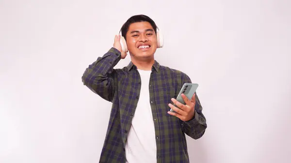 Akıllı telefondan müzik dinleyen Asyalı genç adam beyaz arka planda kablosuz kulaklık kullanıyor.