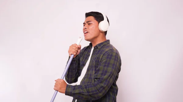 Asyalı adam kulaklıkla müzik dinliyor ve süpürgeyle beyaz arka planda şarkı söylüyor.