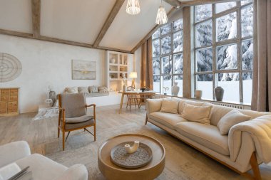 Kış ormanlarına bakan kocaman panoramik pencereli şık bir dağ evinin içi sıcacık. Açık plan, ahşap dekorasyon, sıcak renkler ve aile ocağı.