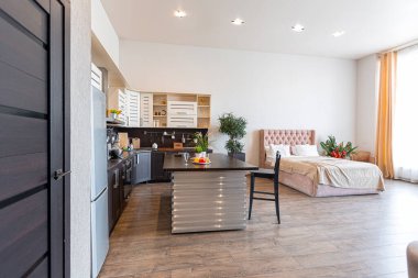 Yumuşak ve sıcak renklerde çağdaş, sıradan bir stüdyo tasarımı. Basit mobilyalar. Güneş ışığıyla aydınlık geniş bir oda. Gündüz vakti. Döşeme kahverengi zemin ve beyaz duvarlar .