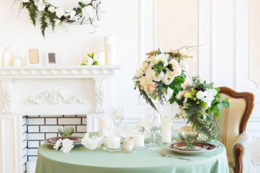 Oturma odasının ışıl ışıl, lüks bir tasarımı var. Süslerle süslenmiş beyaz duvarlar. Şömine. İçeriden kimse yok. Tabakların, mumların ve çiçek buketlerinin olduğu masa..