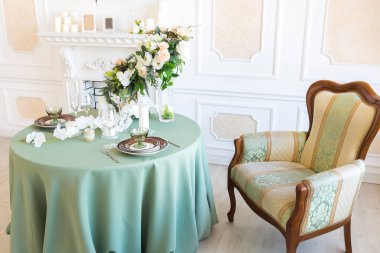 Oturma odasının ışıl ışıl, lüks bir tasarımı var. Süslerle süslenmiş beyaz duvarlar. Şömine. İçeriden kimse yok. Tabakların, mumların ve çiçek buketlerinin olduğu masa..