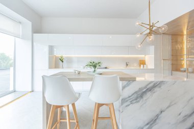 Modern moda kar beyazı mutfağının lüks iç tasarımı. Minimalist tarzda, ada ve iki bar taburesi. Yere kocaman pencereler ve bulaşıklar için cam askı.