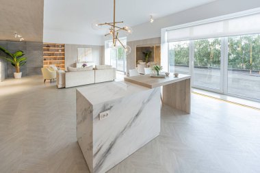 Beyaz ve bej renkli geniş tabandan tavana kadar pencereleri olan büyük bir stüdyonun modern iç tasarımı. Adayı ve dinlenme alanı olan beyaz mutfak.