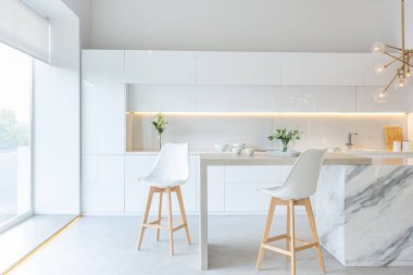 Modern moda kar beyazı mutfağının lüks iç tasarımı. Minimalist tarzda, ada ve iki bar taburesi. Yere kocaman pencereler ve bulaşıklar için cam askı.