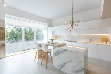 Modern moda kar beyazı mutfağının lüks iç tasarımı. Minimalist tarzda, ada ve iki bar taburesi. Yere kocaman pencereler ve bulaşıklar için cam askı.