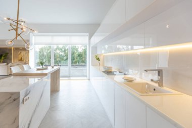 Modern moda kar beyazı mutfağının lüks iç tasarımı. Minimalist tarzda, ada ve iki bar taburesi. Yere kocaman pencereler ve bulaşıklar için cam askı.
