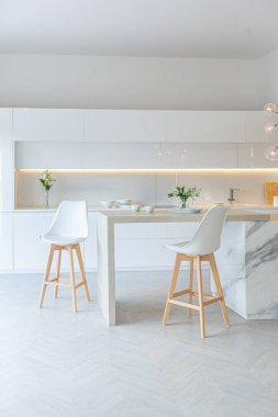 Modern moda kar beyazı mutfağının lüks iç tasarımı. Minimalist tarzda, ada ve iki bar taburesi. Yere kocaman pencereler ve bulaşıklar için cam askı.