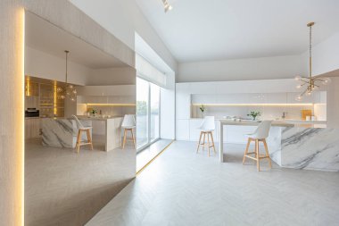Modern moda kar beyazı mutfağının lüks iç tasarımı. Minimalist tarzda, ada ve iki bar taburesi. Yere kocaman pencereler ve bulaşıklar için cam askı.