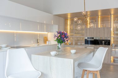 Modern moda kar beyazı mutfağının lüks iç tasarımı. Minimalist tarzda, ada ve iki bar taburesi. Yere kocaman pencereler ve bulaşıklar için cam askı.