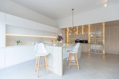 Modern moda kar beyazı mutfağının lüks iç tasarımı. Minimalist tarzda, ada ve iki bar taburesi. Yere kocaman pencereler ve bulaşıklar için cam askı.