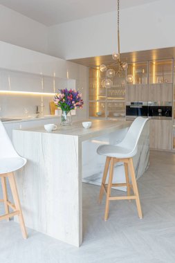 Modern moda kar beyazı mutfağının lüks iç tasarımı. Minimalist tarzda, ada ve iki bar taburesi. Yere kocaman pencereler ve bulaşıklar için cam askı.