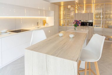 Modern moda kar beyazı mutfağının lüks iç tasarımı. Minimalist tarzda, ada ve iki bar taburesi. Yere kocaman pencereler ve bulaşıklar için cam askı.