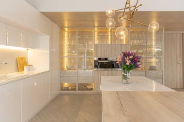 Modern moda kar beyazı mutfağının lüks iç tasarımı. Minimalist tarzda, ada ve iki bar taburesi. Yere kocaman pencereler ve bulaşıklar için cam askı.