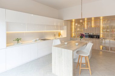 Modern moda kar beyazı mutfağının lüks iç tasarımı. Minimalist tarzda, ada ve iki bar taburesi. Yere kocaman pencereler ve bulaşıklar için cam askı.