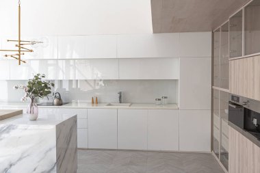Modern moda kar beyazı mutfağının lüks iç tasarımı. Minimalist tarzda, ada ve iki bar taburesi. Yere kocaman pencereler ve bulaşıklar için cam askı.