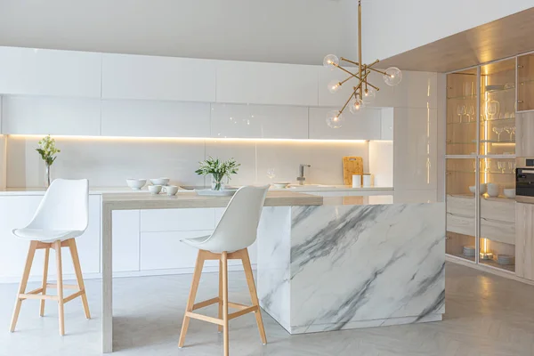 Modern moda kar beyazı mutfağının lüks iç tasarımı. Minimalist tarzda, ada ve iki bar taburesi. Yere kocaman pencereler ve bulaşıklar için cam askı.