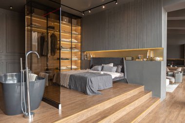 Yatak odası ve cam bölmenin arkasında özgür bir banyo lüks bir evin şık bir iç mekanında, koyu modern tasarımla ahşap süslemeli ve yol gösterici aydınlatmalı