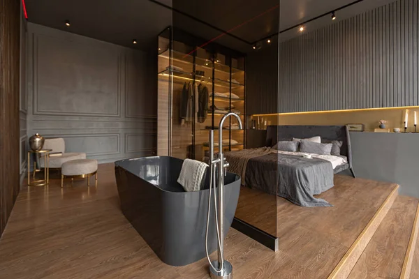 木製のトリムと光を導いたダークモダンデザインの高級住宅のシックな高価なインテリアのガラスのパーティションの後ろのベッドルームと自立風呂 — ストック写真