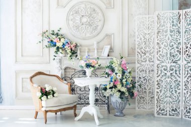 Parlak, beyaz ve mavi renkli, vazolarında çiçekler olan bir oturma odası. Duvarlar barok süslemelerle süslenmiş.