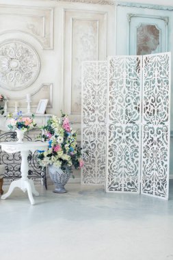 Parlak, beyaz ve mavi renkli, vazolarında çiçekler olan bir oturma odası. Duvarlar barok süslemelerle süslenmiş.