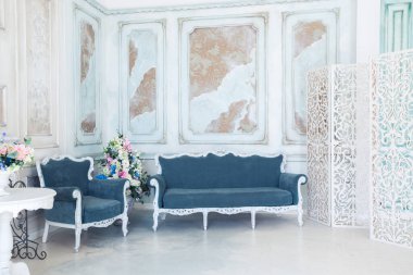 Parlak, beyaz ve mavi renkli, vazolarında çiçekler olan bir oturma odası. Duvarlar barok süslemelerle süslenmiş.