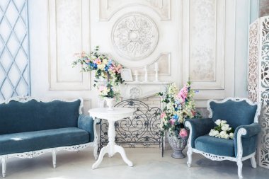 Parlak, beyaz ve mavi renkli, vazolarında çiçekler olan bir oturma odası. Duvarlar barok süslemelerle süslenmiş.