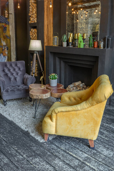dark brutal interior of sitting room decorated with wooden logs. yellow and grey soft armchairs, huge arc window and fireplace