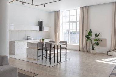 Güneşli bir günde pahalı bir evin minimalist modern lüks tasarımında yüksek bar tabureleri olan mutfak ve yemek adası. Alçılı beyaz duvarlar, parke, dekoratif ışıklandırma ve içeride kimse yok.