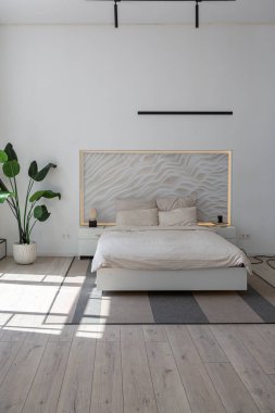 Güneşli bir günde pahalı bir evin minimalist modern lüks tasarımı için yatak odası. Yatak başlığında dekoratif panel, alçılı beyaz duvarlar, parke, dekoratif ışıklandırma ve hiç kimse