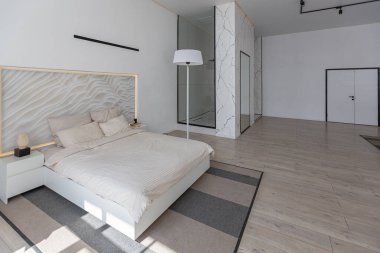 Güneşli bir günde pahalı bir evin minimalist modern lüks tasarımı için yatak odası. Yatak başlığında dekoratif panel, alçılı beyaz duvarlar, parke, dekoratif ışıklandırma ve hiç kimse