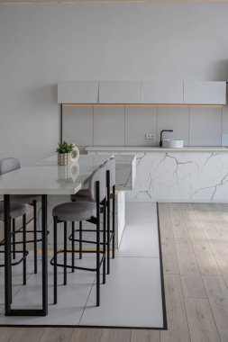 Güneşli bir günde pahalı bir evin minimalist modern lüks tasarımında yüksek bar tabureleri olan mutfak ve yemek adası. Alçılı beyaz duvarlar, parke, dekoratif ışıklandırma ve içeride kimse yok.