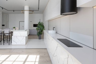 Güneşli bir günde pahalı bir evin minimalist modern lüks tasarımında yüksek bar tabureleri olan mutfak ve yemek adası. Alçılı beyaz duvarlar, parke, dekoratif ışıklandırma ve içeride kimse yok.