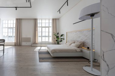 Güneşli bir günde pahalı bir evin minimalist modern lüks tasarımı için yatak odası. Yatak başlığında dekoratif panel, alçılı beyaz duvarlar, parke, dekoratif ışıklandırma ve hiç kimse