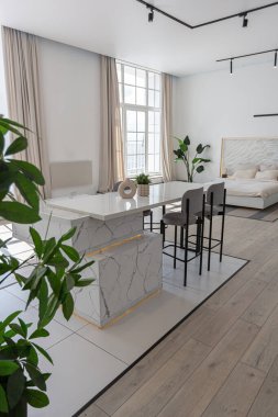 Güneşli bir günde pahalı bir evin minimalist modern lüks tasarımında yüksek bar tabureleri olan mutfak ve yemek adası. Alçılı beyaz duvarlar, parke, dekoratif ışıklandırma ve içeride kimse yok.