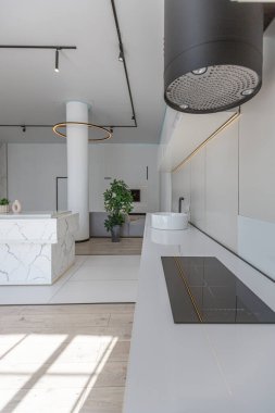 Güneşli bir günde pahalı bir evin minimalist modern lüks tasarımında yüksek bar tabureleri olan mutfak ve yemek adası. Alçılı beyaz duvarlar, parke, dekoratif ışıklandırma ve içeride kimse yok.