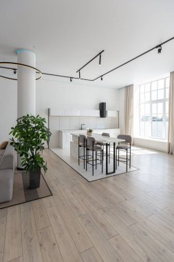 Güneşli bir günde pahalı bir evin minimalist modern lüks tasarımında yüksek bar tabureleri olan mutfak ve yemek adası. Alçılı beyaz duvarlar, parke, dekoratif ışıklandırma ve içeride kimse yok.