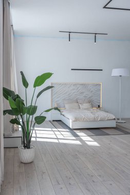 Güneşli bir günde pahalı bir evin minimalist modern lüks tasarımı için yatak odası. Yatak başlığında dekoratif panel, alçılı beyaz duvarlar, parke, dekoratif ışıklandırma ve hiç kimse