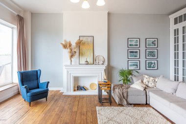 Geniş, modern, çok parlak, pastel renkli oturma odası, gri kanepe, klasik şömine ve ahşap zemin.