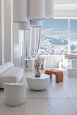Modern, saf, beyaz bir iç tasarım. Yumuşak stil mobilyaları olan şık bir oturma odası. Geniş pencere çerçevesi gün ışığında sakin bir atmosfer yaratıyor.