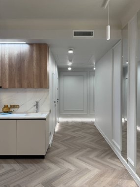 Çağdaş bir apartman dairesi, parlak ahşap döşemeleri ve modern aydınlatma armatürleriyle geniş bir yerleşim alanı sergiliyor. Mutfak alanında temiz bir görünüm için minimalist dolaplar var..