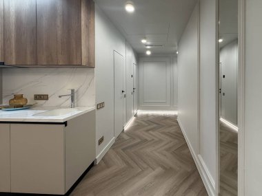 Çağdaş bir apartman dairesi, parlak ahşap döşemeleri ve modern aydınlatma armatürleriyle geniş bir yerleşim alanı sergiliyor. Mutfak alanında temiz bir görünüm için minimalist dolaplar var..