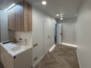 Çağdaş bir apartman dairesi, parlak ahşap döşemeleri ve modern aydınlatma armatürleriyle geniş bir yerleşim alanı sergiliyor. Mutfak alanında temiz bir görünüm için minimalist dolaplar var..
